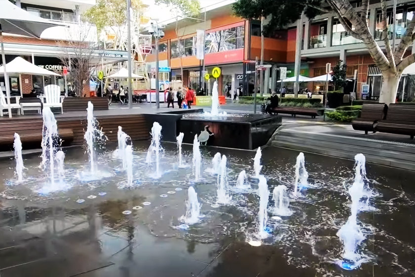 Rouse Hill Town Centre open-air plaza with ground-level water fountain feature, The Coffee Club cafe, timber bench seating and colourful retail facades
